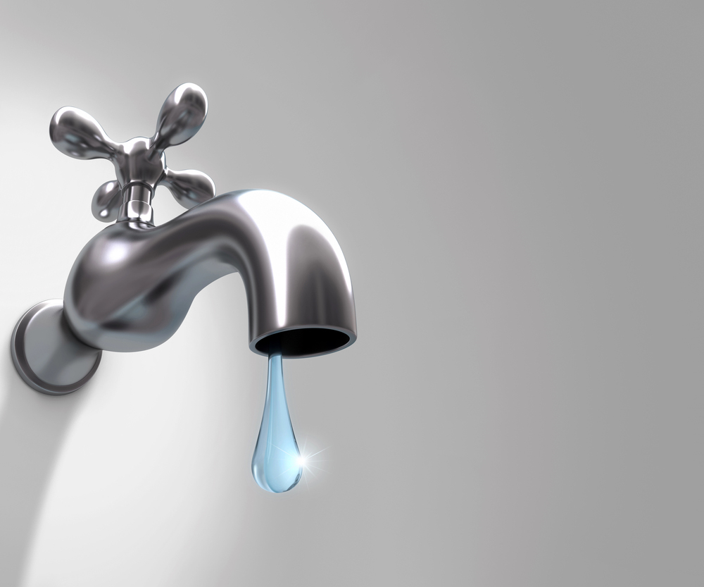 A shiny metal tap mounted on a plain wall releases a single, clear drop of water. The background is a light grey gradient, and the drop catches the light as it falls.