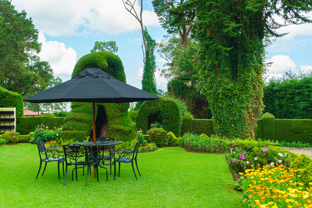 A black metal table with four chairs sits under a large black umbrella in a lush garden with green hedges, trimmed topiary, and colourful flowers on a sunny day.