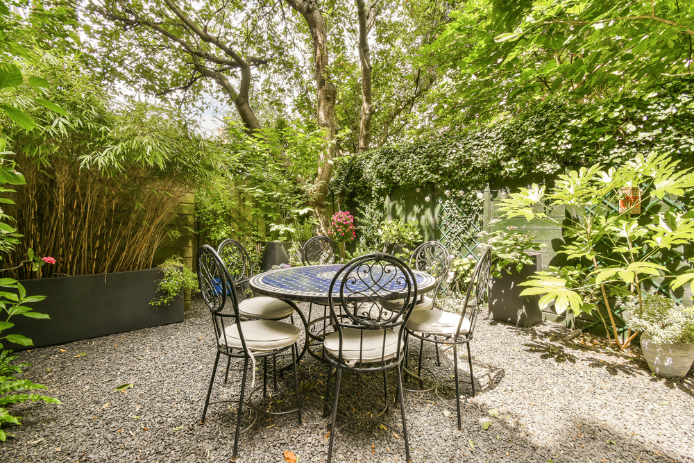 A round table with six chairs sits on gravel in a lush garden surrounded by greenery, tall plants, and trees, creating a peaceful outdoor dining area.