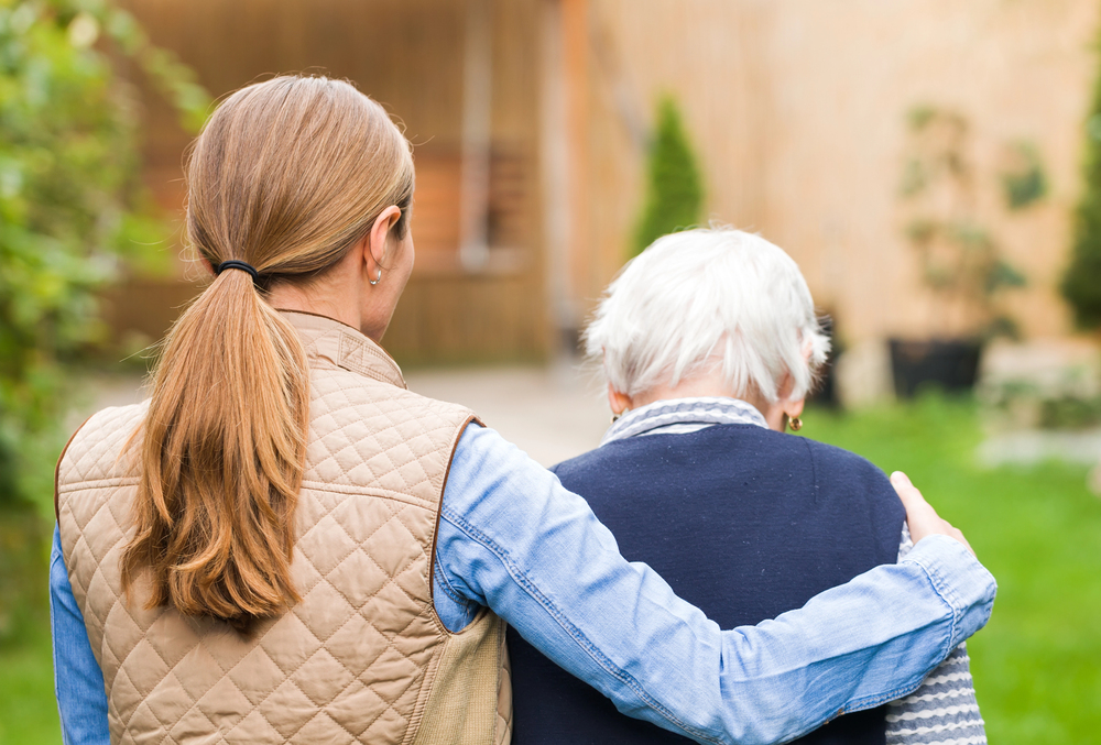 A younger woman with a ponytail has her arm round an older woman with white hair as they walk outside in a garden, seen from behind.