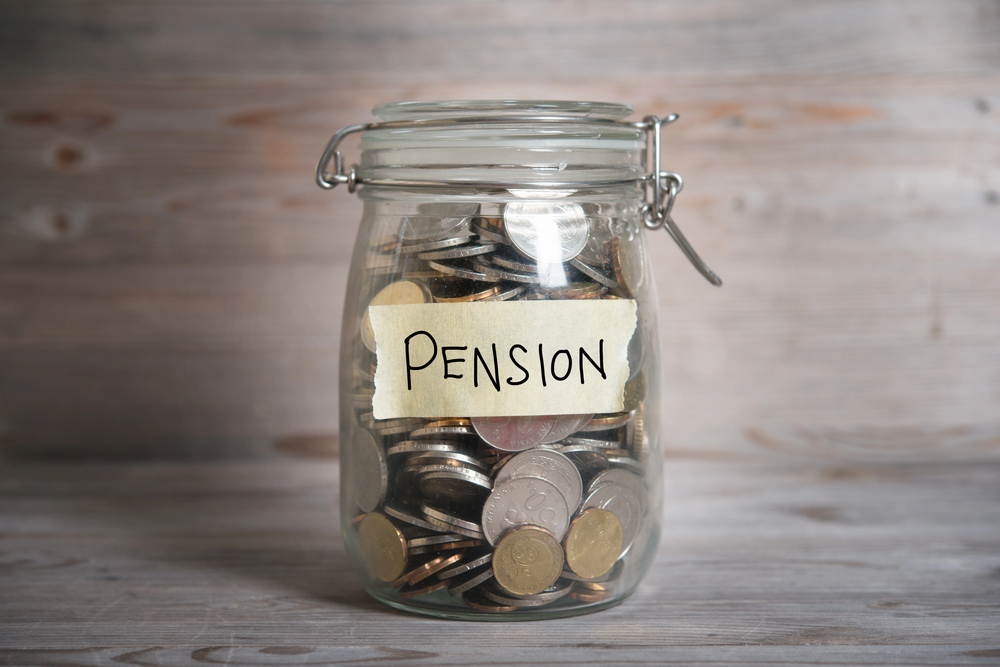 A glass jar labelled PENSION filled with various coins, sitting on a wooden surface with a light brown, wooden background.