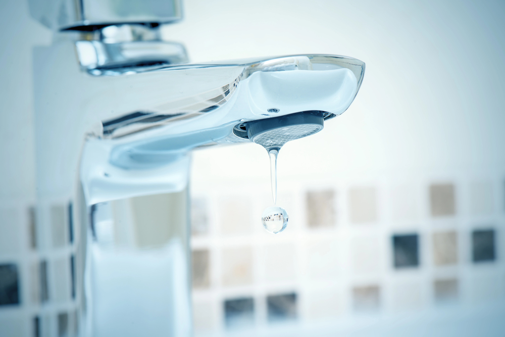 A close-up of a chrome bathroom tap with a single water droplet hanging from it. The background shows a tiled wall in neutral colours, and the scene suggests a leaky tap.