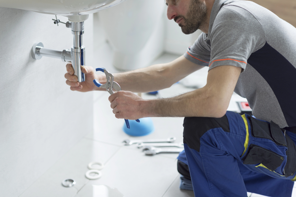 A plumber kneels on the floor fixing a pipe under a sink using a spanner. Various tools and plumbing parts are scattered nearby.