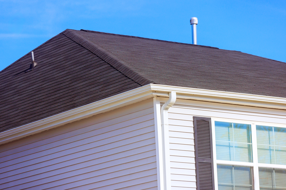 A close-up of a house's upper corner shows brown roof tiles, beige cladding, white gutters, a metal vent pipe, and part of a window with brown shutters against a clear blue sky.