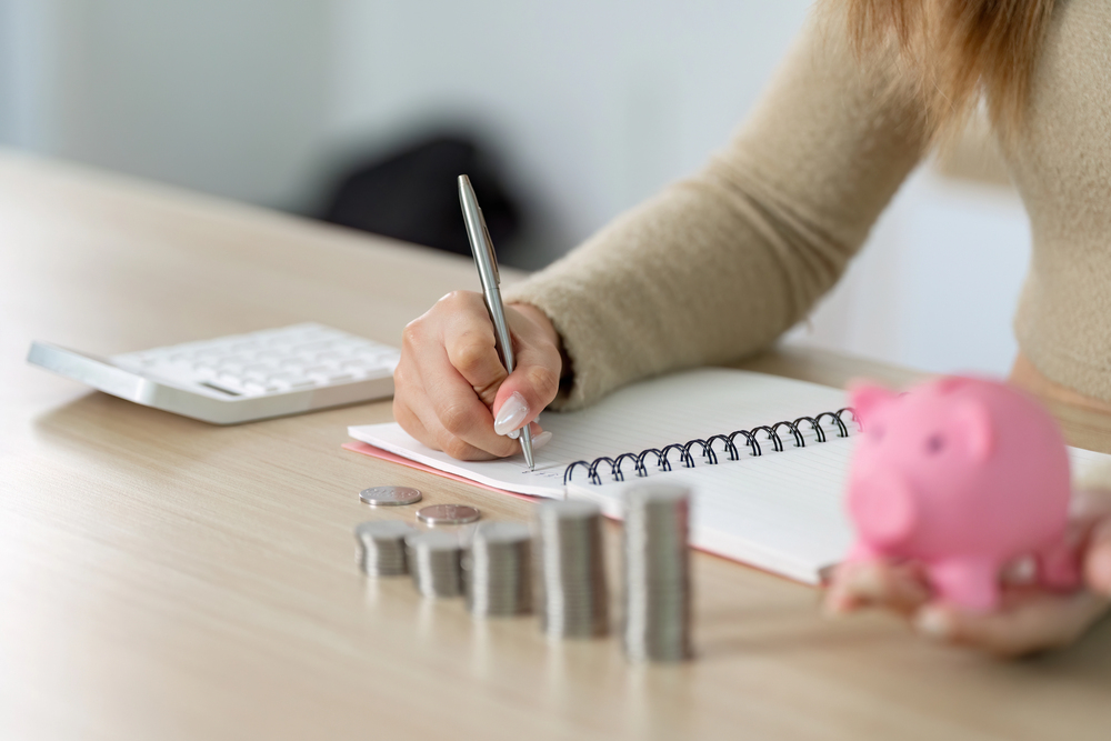 A person writes in a notebook at a desk with a calculator, a pink piggy bank, and neatly stacked coins in the foreground, symbolising budgeting or financial planning.