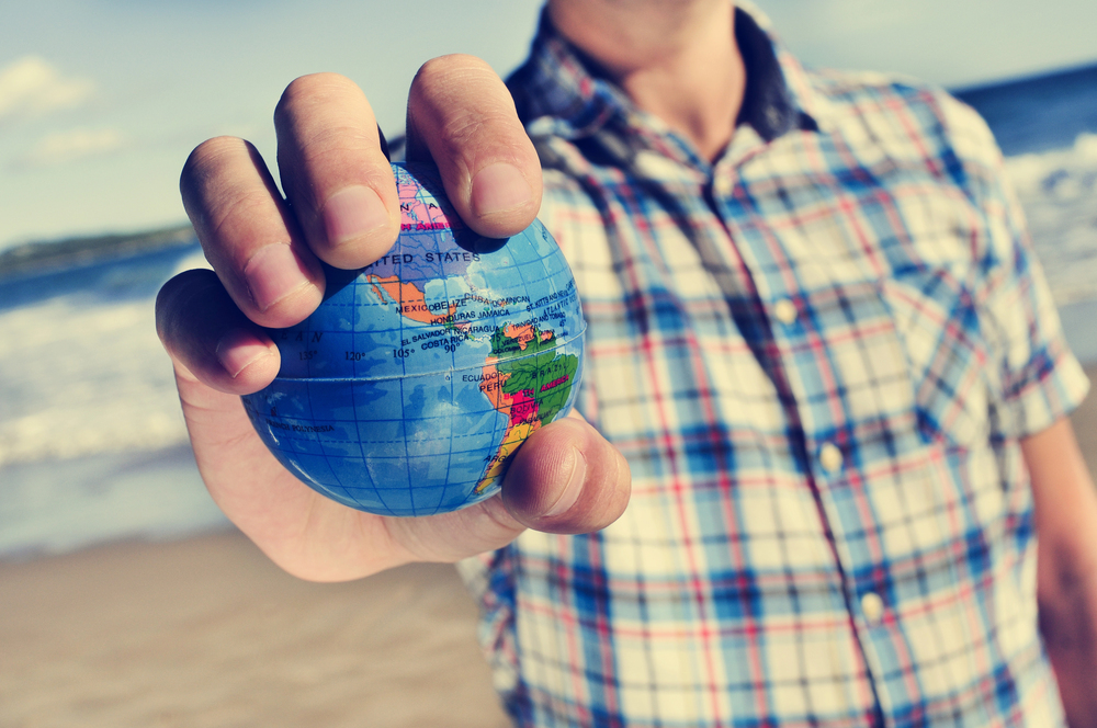 A person in a checked shirt holds a small globe in their hand on a beach, with the sea and sky blurred in the background.