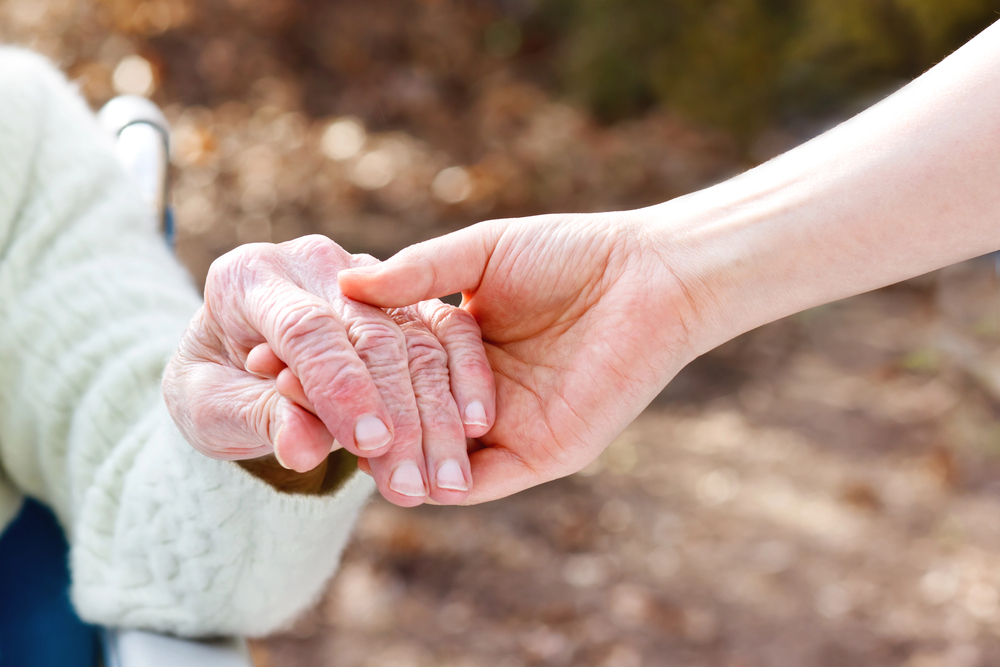 A younger person gently holds the hand of an elderly person, offering comfort and support. The elderly person wears a light jumper. The background is softly blurred, focussing on the hands.
