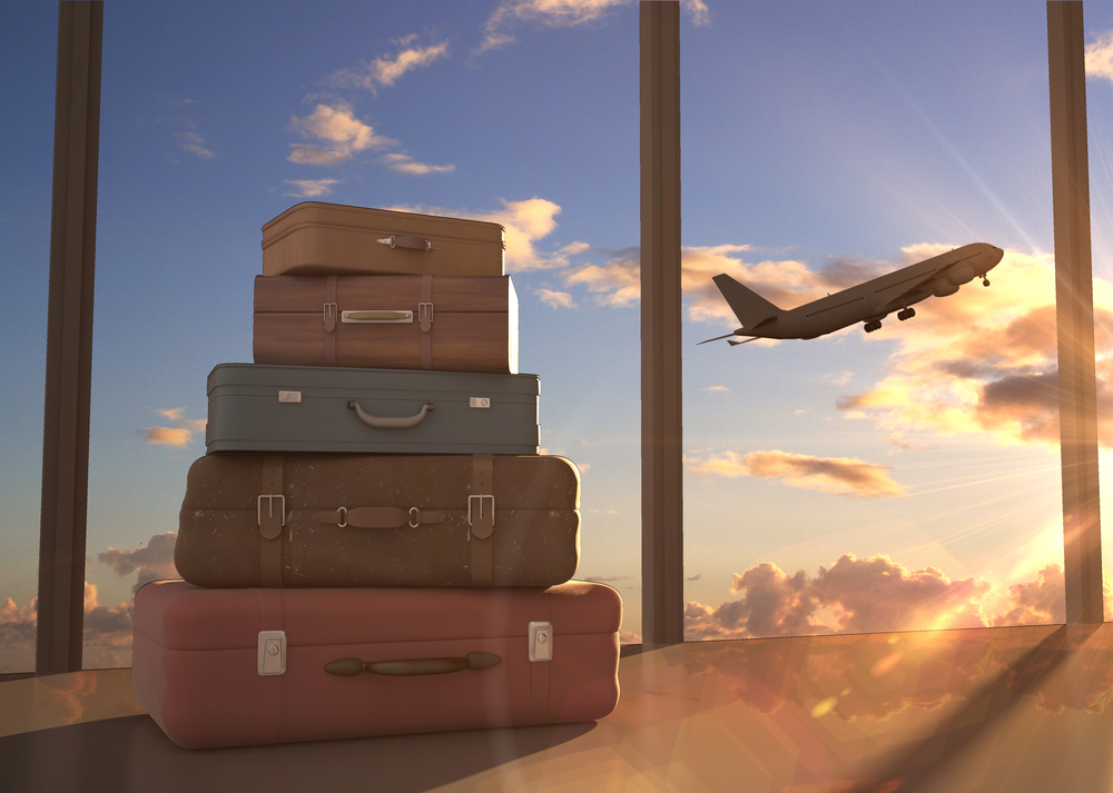 A stack of four suitcases sits by large airport windows at sunset, with an aeroplane taking off in the sky outside.