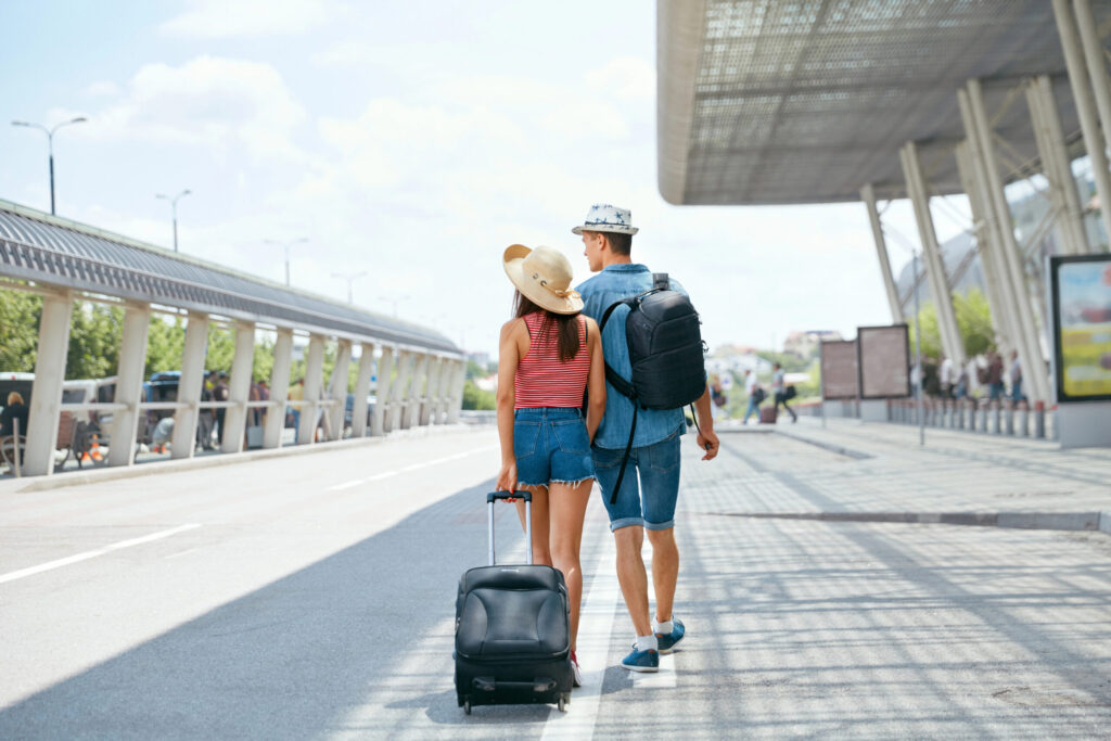 A couple walks away from the camera down an outdoor walkway, each carrying a rucksack; the woman also pulls a suitcase. Both wear summer clothes and hats, with a modern building structure visible to their right.