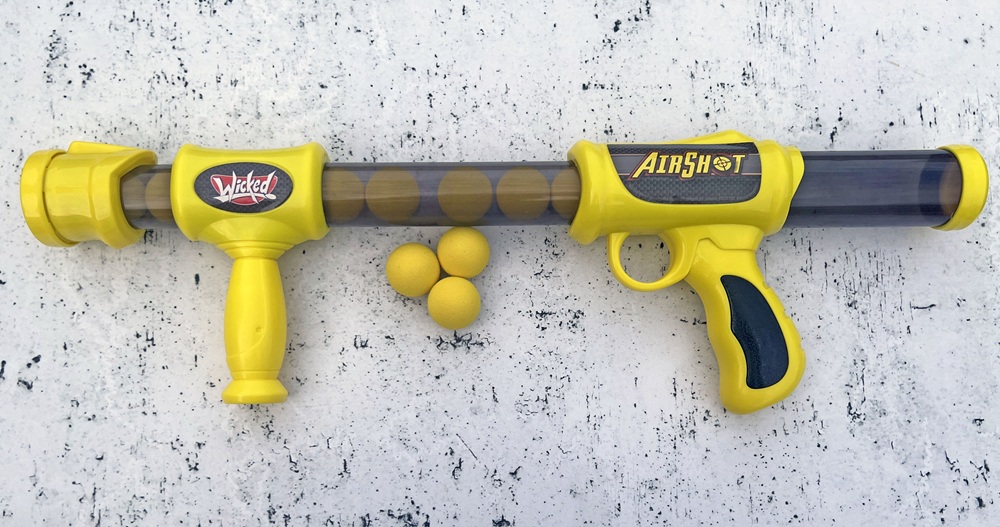 A yellow plastic toy gun called Airshot with a handle and barrel, beside four small yellow foam balls, on a white speckled surface.