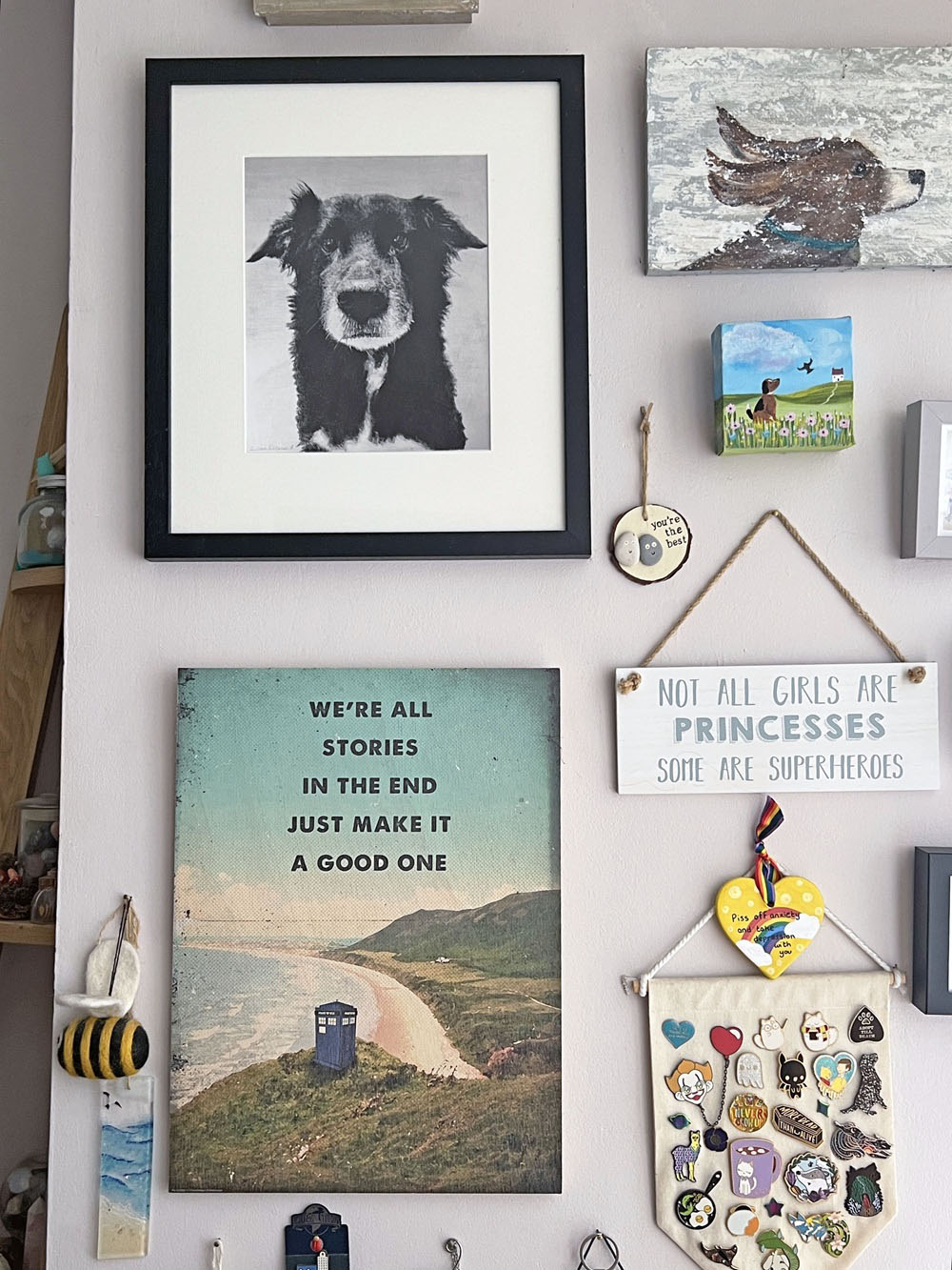 A collage wall with dog artwork, a bee ornament, a Dr Who landscape poster, a sign about princesses and superheroes, and various small decorative items including badges and painted pieces.
