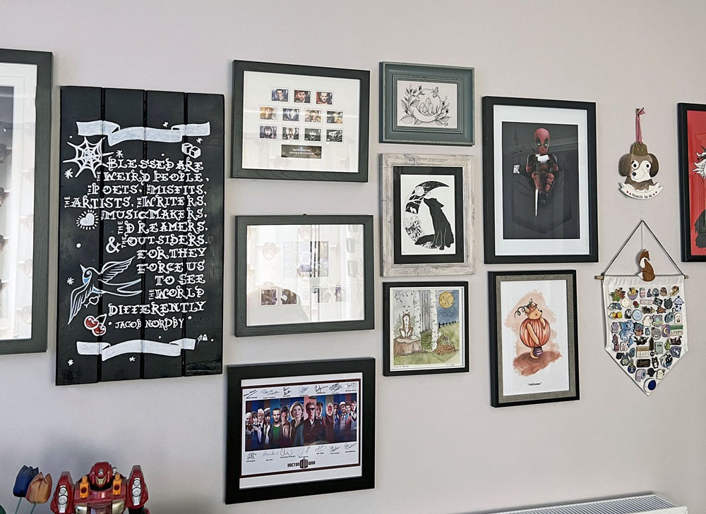Gallery wall with framed art, quotes, illustrations, and memorabilia, including a hanging plaque and Iron Man figure at the bottom left. The arrangement features various sizes and styles on a light-coloured wall.