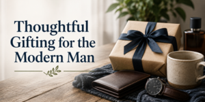 Gift box with a black ribbon, wallet, watch, and mug on a table. Text reads: Thoughtful Gifting for the Modern Man. Greenery and a cologne bottle are also visible.