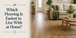 Living room with wood-look flooring, beige sofa with cushions, wooden coffee table on a woven rug, plants, and text: Which Flooring Is Easiest to Live With at Home? on a cream background.