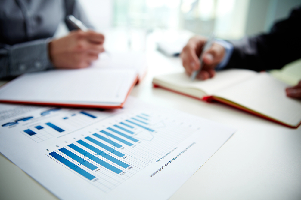 Two people writing in notebooks at a desk with a printed bar graph and data chart in the foreground, suggesting a business meeting or financial discussion.