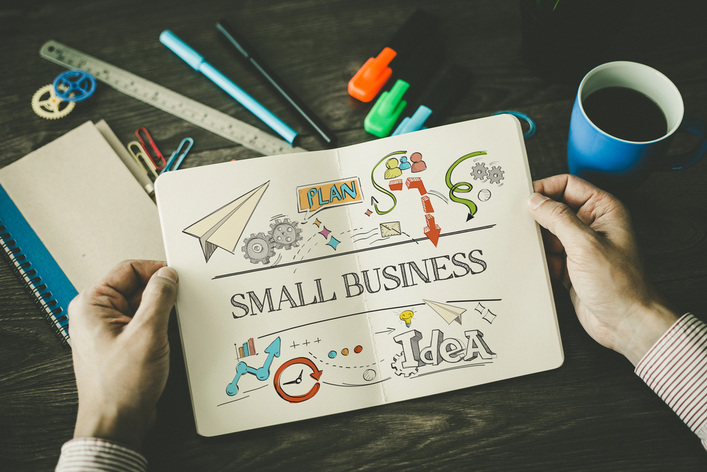 Person holding a notebook open to colourful sketches and words: Plan, Small Business, and Idea. Office supplies, documents, and a cup of coffee are on a dark desk in the background.