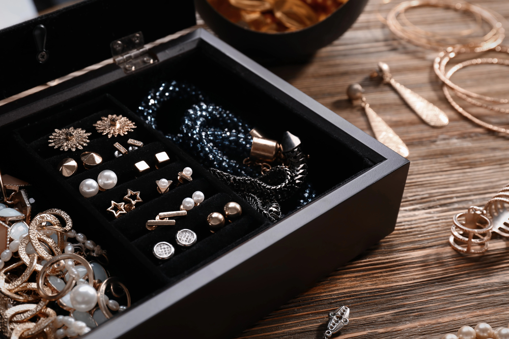 An open black jewellery box filled with assorted earrings, rings, and necklaces sits on a wooden surface, with more jewellery items scattered around it.