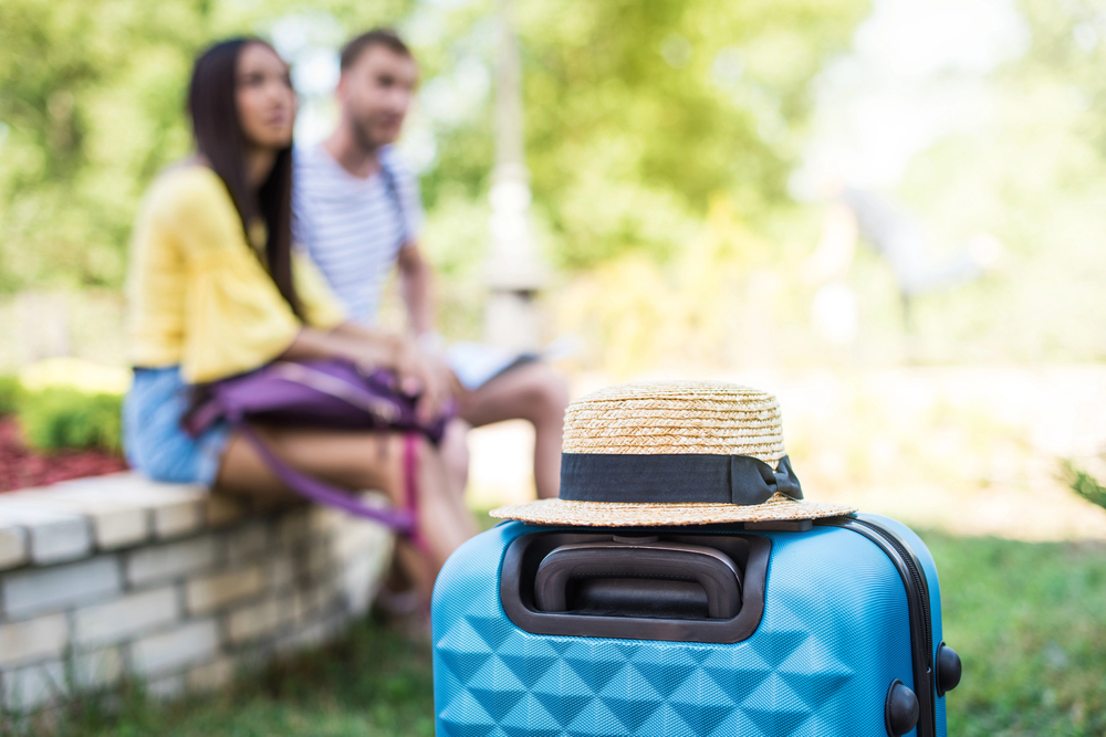 A straw hat sits on a blue suitcase in focus, whilst a man and a woman sit together on a stone bench in the blurred background outdoors.