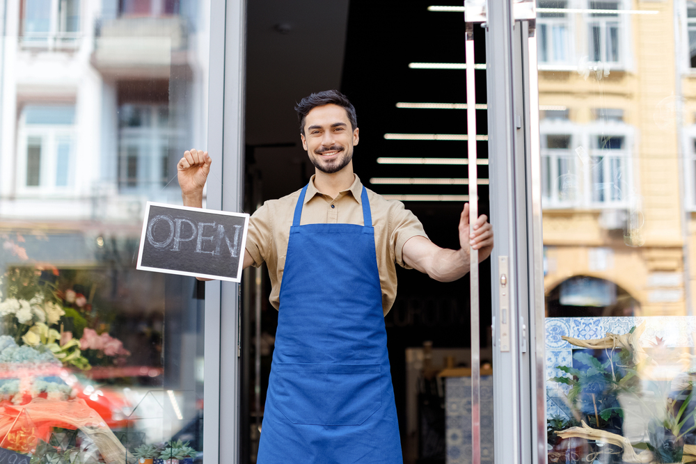 A smiling person wearing a blue apron stands in a shop doorway, holding an OPEN sign, welcoming customers inside.