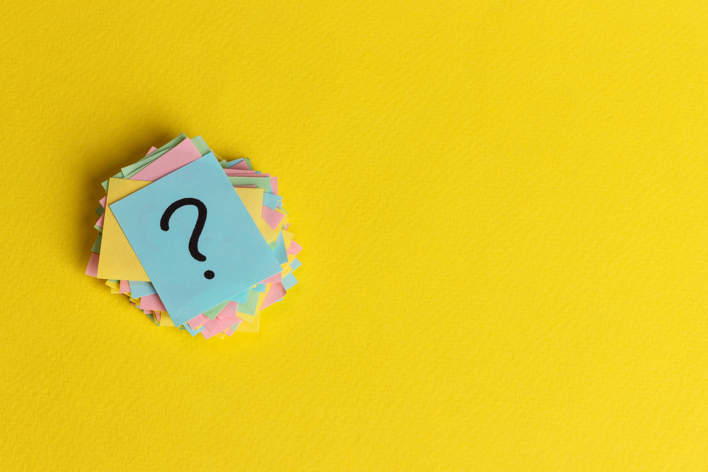 A stack of pastel-coloured sticky notes with a black question mark on the top note, set on a bright yellow background.