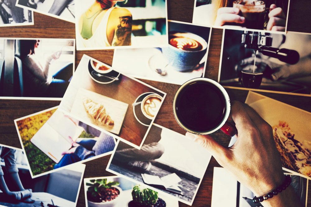 A hand holding a cup of coffee over a table scattered with colourful photographs of people, coffee, food, and plants.