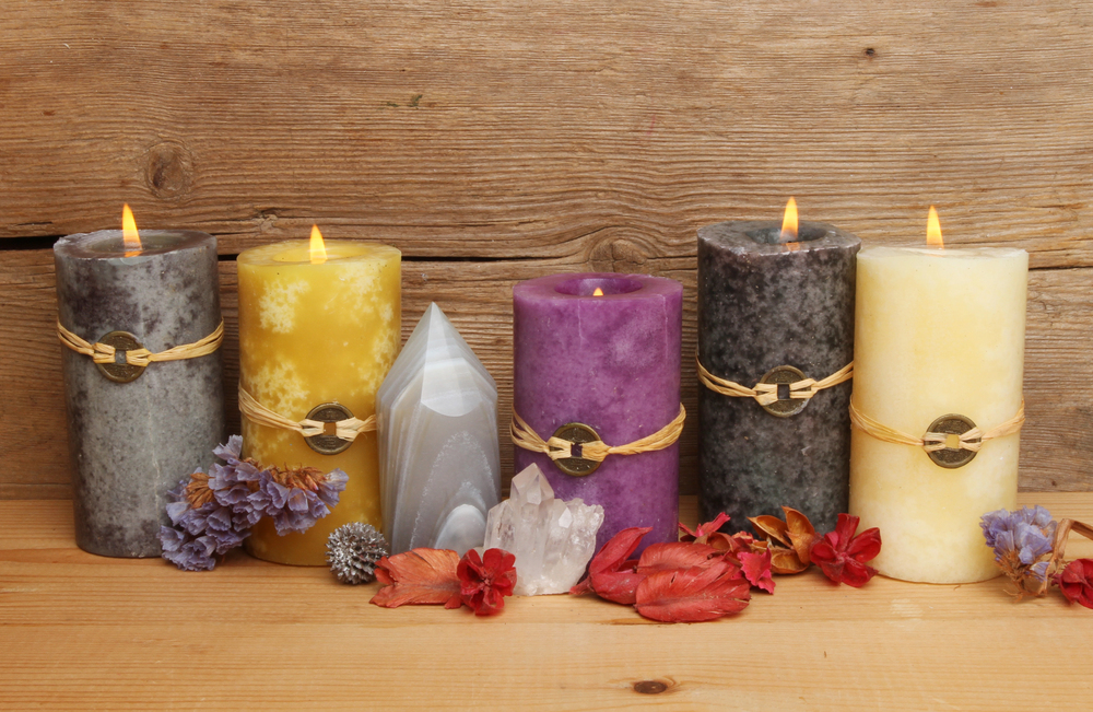 Five lit pillar candles, in grey, yellow, purple, and cream colours, sit on a wooden surface among dried flowers and crystals, against a wood background.