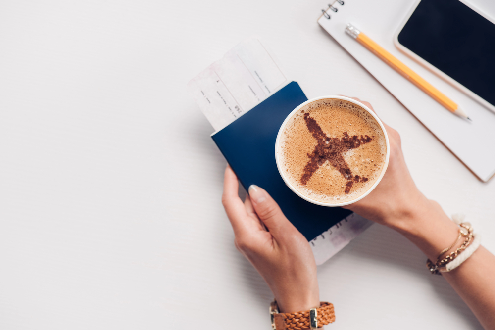 Hands holding a cup of coffee with an aeroplane design in the froth, a passport and boarding pass. Nearby are a notepad, pencil, and a mobile phone on a white surface.