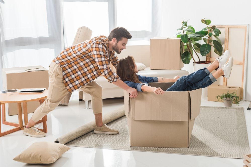 A man playfully pushes a woman sitting in a cardboard box amid moving boxes and furniture in a bright lounge.