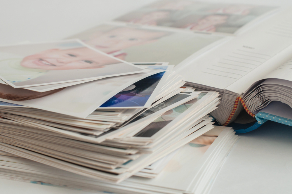 A stack of printed photographs lies next to an open photo album filled with more pictures, some showing close-ups of children's faces, all arranged on a light-coloured surface.