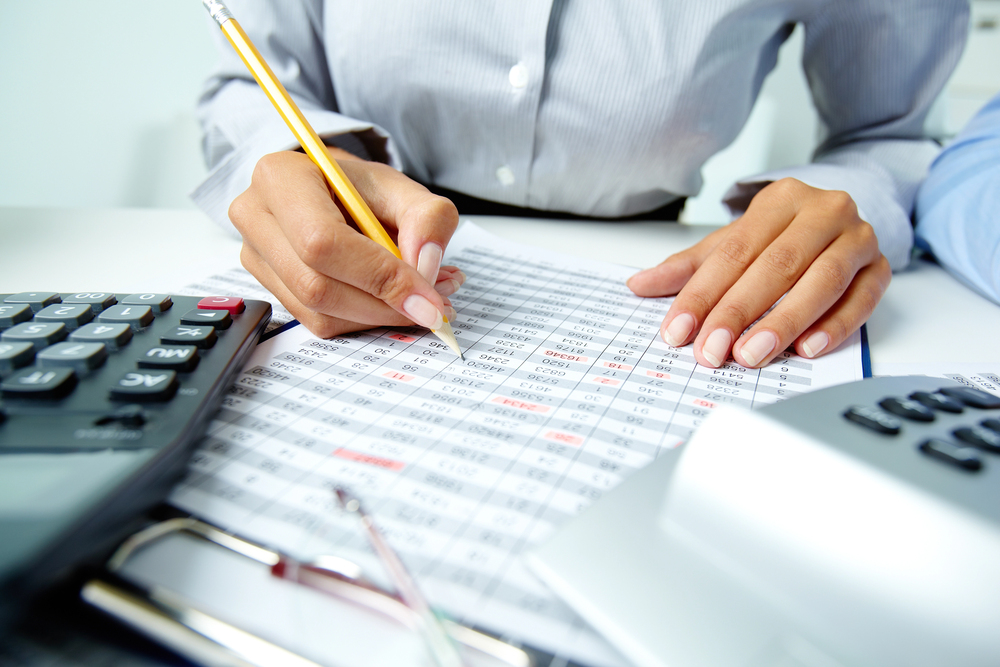 Close-up of a person's hands working with a spreadsheet, using a pencil. Calculators, documents, and a clipboard are also visible on the desk. The person is wearing a striped shirt.