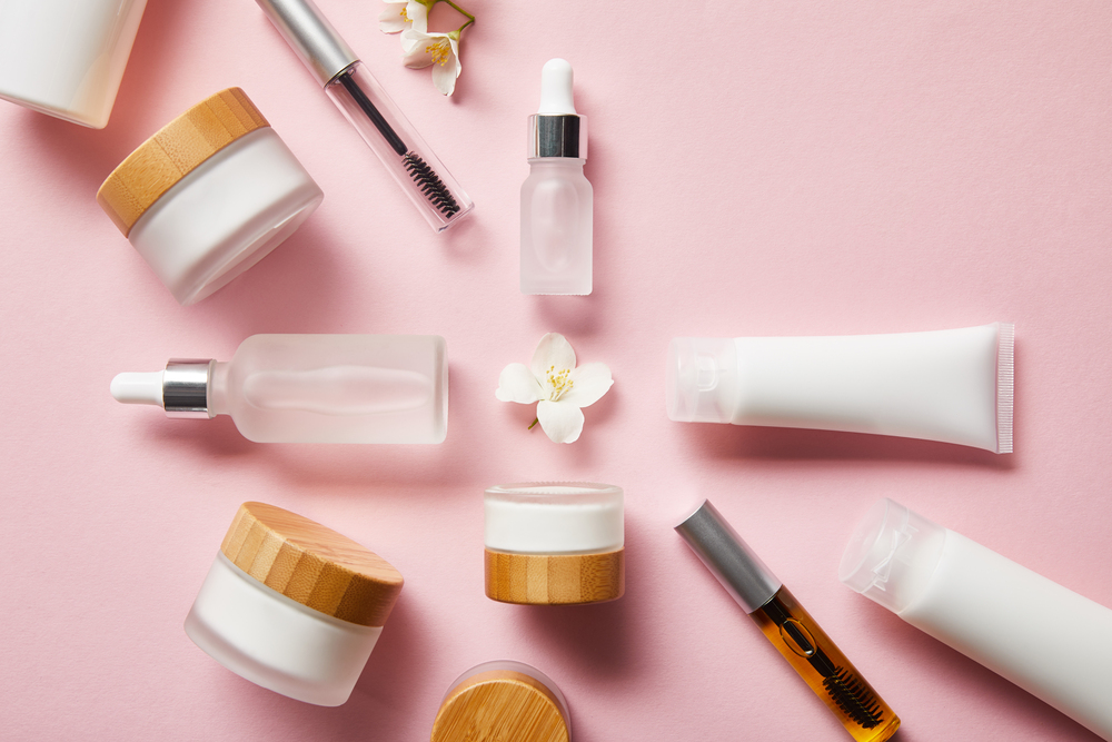 Assorted skincare containers—jars, tubes, dropper bottles, and mascara wands—arranged in a circle on a pink background with small white flowers.