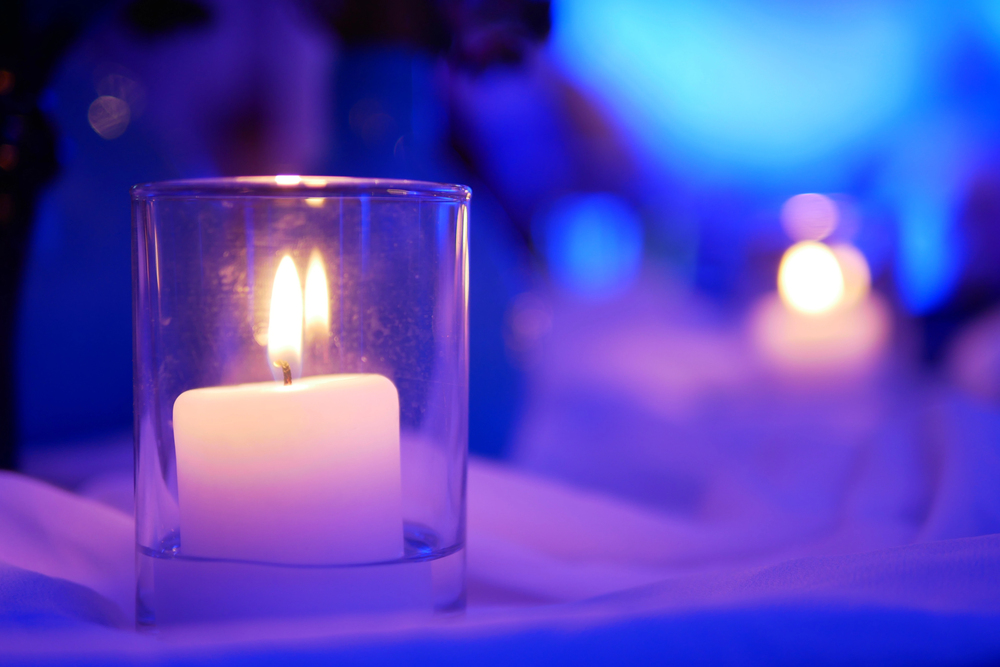 A white candle burns inside a clear glass holder, with another blurred candle glowing in the blue-lit background.
