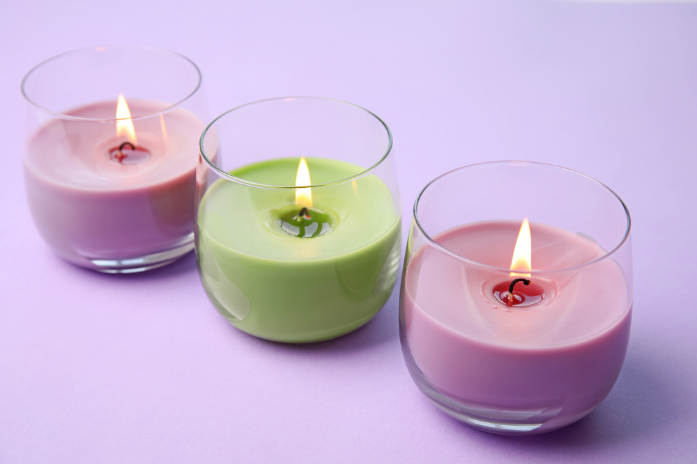 Three lit candles in round glass holders sit on a light purple surface. The candles are pastel purple, green, and pink, and each has a visible flame.