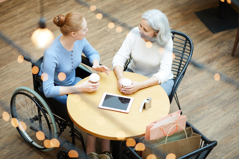 Two women sit at a round table in a café, smiling and talking over coffee. One uses a wheelchair. An electronic tablet, shopping bags, and takeaway containers are on the table. Warm lights hang above.