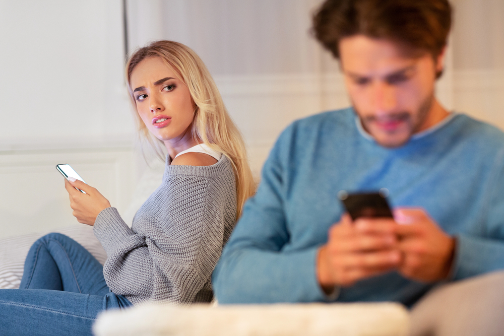 A woman looks suspiciously at a man using his mobile while she holds her own mobile. Both are sitting on a sofa in a brightly lit room.