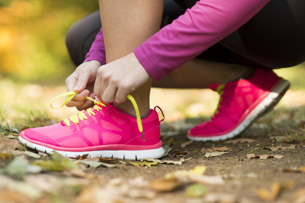 A person wearing a pink long-sleeve top ties the yellow laces of bright pink running trainers while kneeling on a leaf-covered outdoor path.