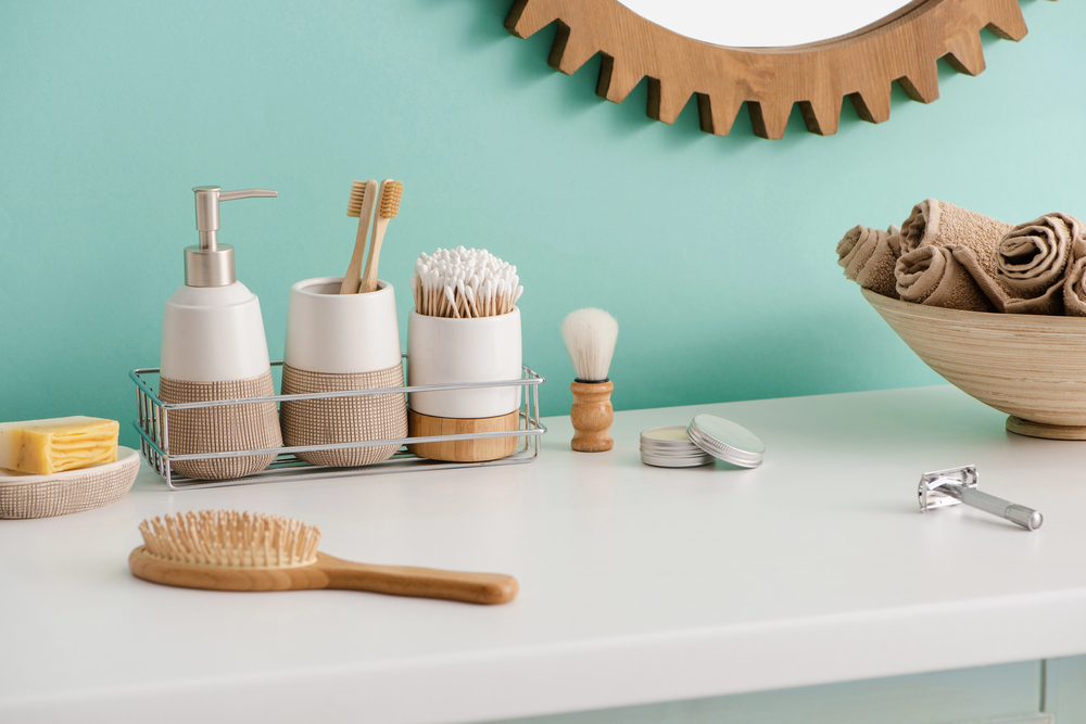 A bathroom countertop with toiletries, including a soap dispenser, toothbrushes, cotton buds, a shaving brush, towel rolls, a wooden hairbrush, razor, and soap, against a turquoise wall and round mirror.