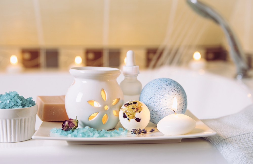A white plate holds a lit candle, bath salts, a soap bar, a blue bath bomb, an oil dropper, and a white ceramic diffuser by a bathtub, with lit candles in the background.