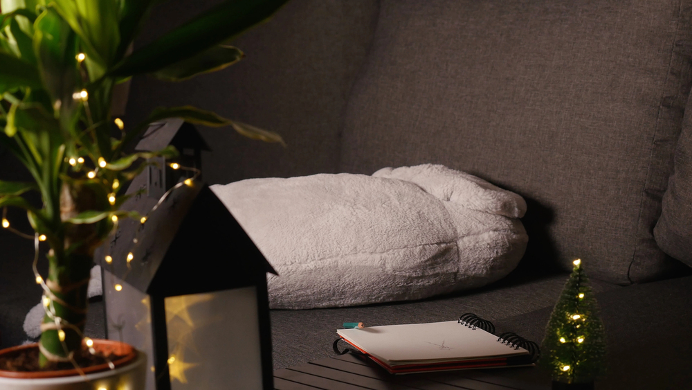 A cosy grey sofa with a white blanket, a potted plant, a black lantern, a small notebook, and a tiny tree wrapped in fairy lights, creating a warm, inviting atmosphere.