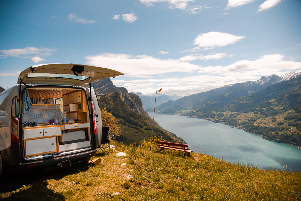 A camper van with its back doors open overlooks a large lake and mountains under a partly cloudy sky. A bench and flag are nearby on the grassy hill.