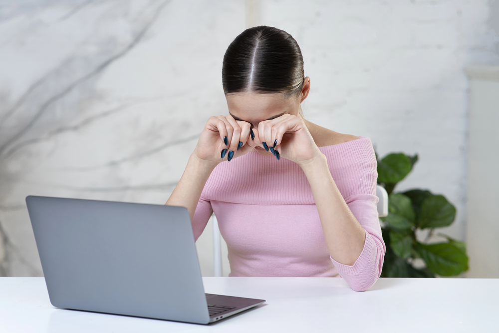 A woman in a pink top sits at a desk with a laptop, rubbing her eyes as if tired or stressed. There is a green plant in the background.