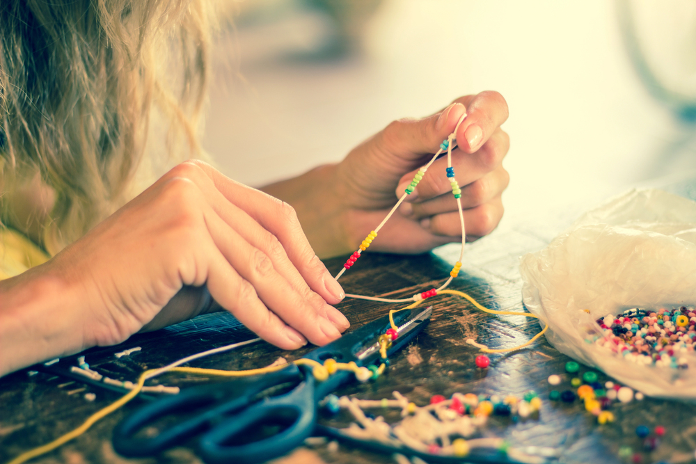 Close-up of a person's hands threading colourful beads onto a cord, with scissors and scattered beads on a wooden table.