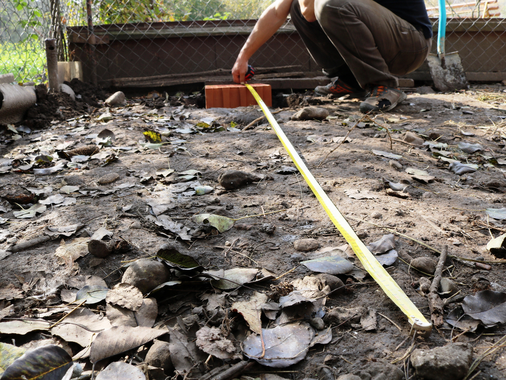 A person squats on the ground using a yellow measuring tape to measure a distance outdoors, surrounded by soil, stones, and dry leaves near a chain-link fence.
