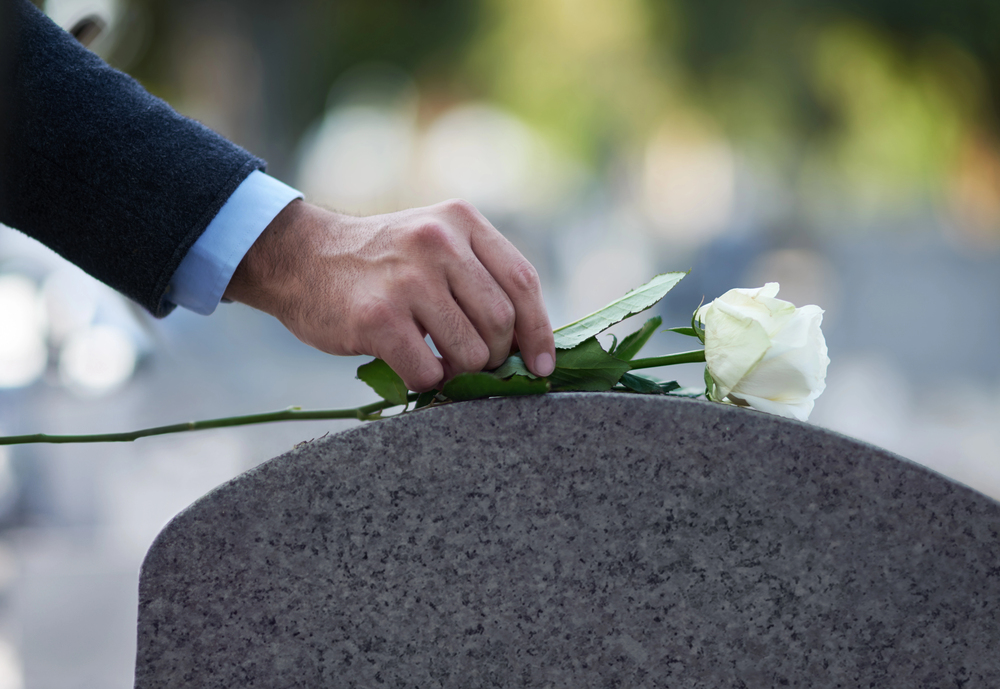 A person's hand gently places a white rose on top of a grey gravestone in a cemetery, symbolising remembrance and mourning.