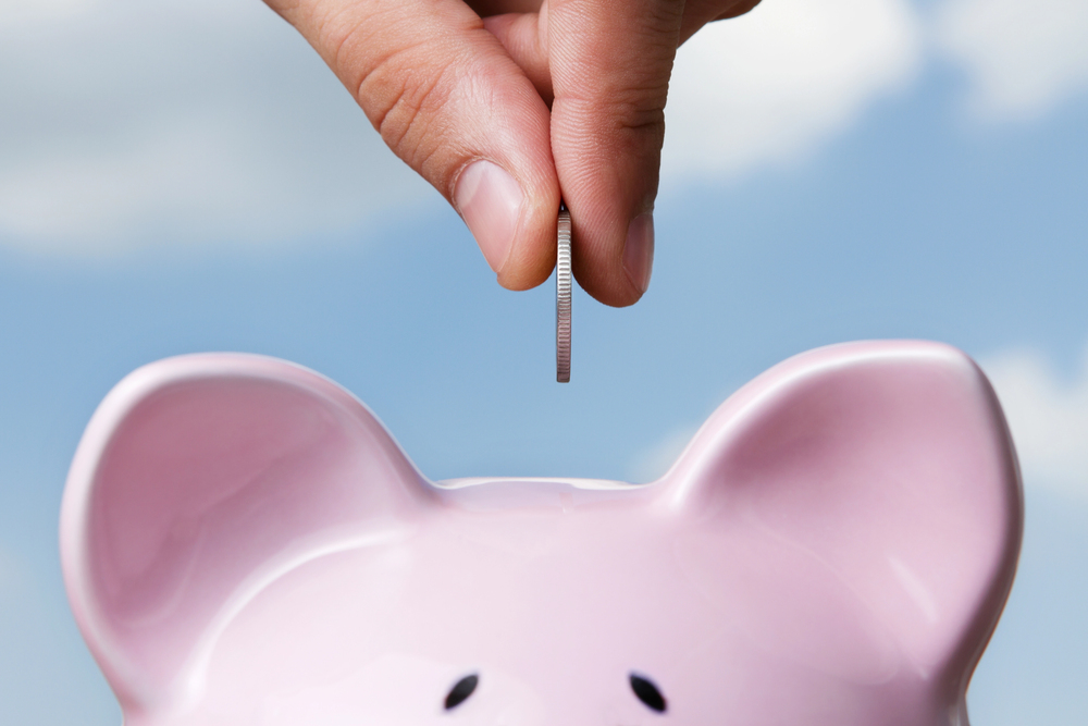 A hand holds a coin above a pink piggy bank against a blue sky, preparing to drop the coin into the slot.