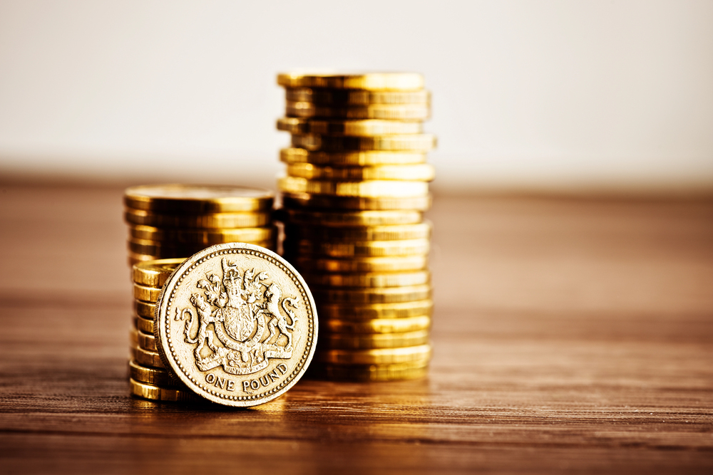 Stacks of British one-pound coins arranged on a wooden surface, with one coin standing upright in front, showing the detailed design on its face.