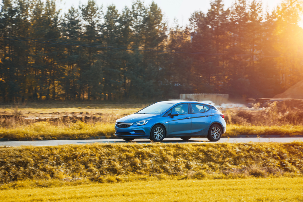 A blue car drives on a road through a rural area with trees and golden sunlight in the background.