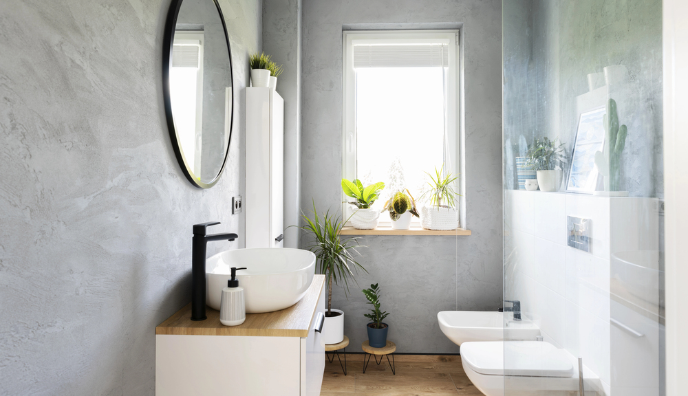 Modern bathroom with a round mirror, white basin, black tap, and wooden countertop. Window with potted plants on the sill lets in natural light. Toilet and bidet are next to a glass shower.