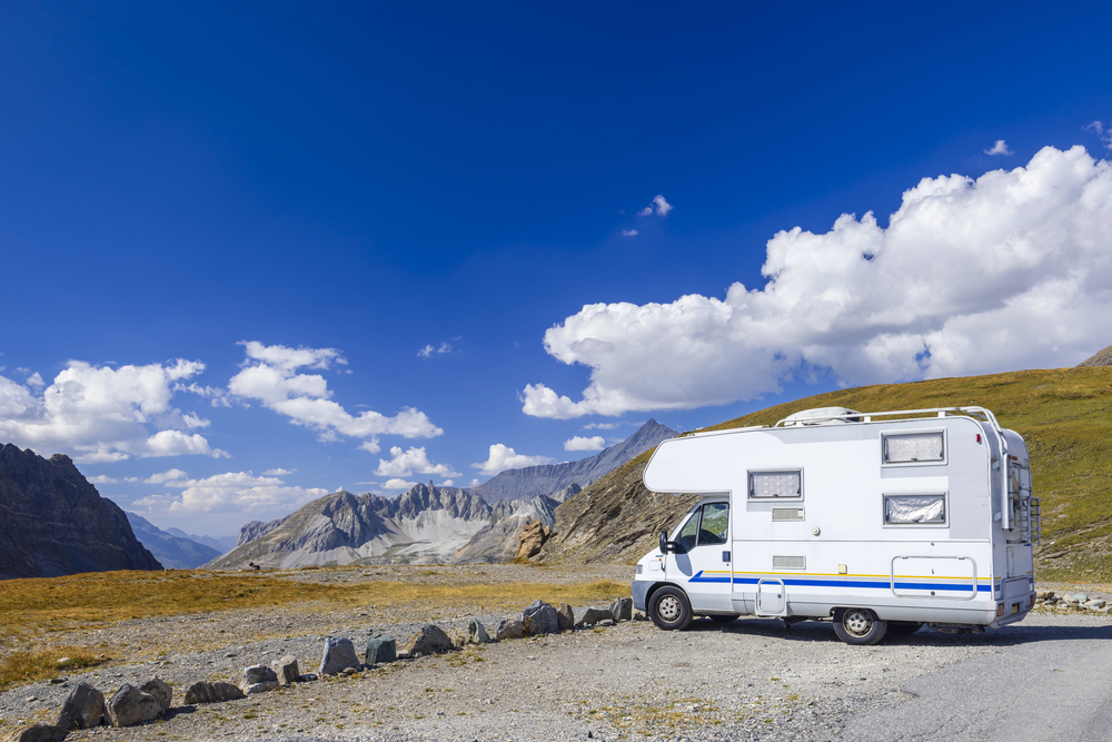 A white camper van is parked on a gravelled area in a mountain landscape under a blue sky with scattered clouds. Rocky peaks and grassy slopes are visible in the background.
