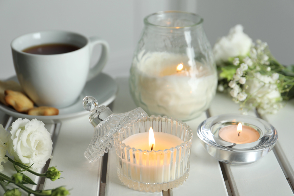 A white table with lit candles, white flowers, a cup of tea on a saucer with biscuits, and a glass candle holder with a lid resting beside it. The scene exudes a calm, cosy atmosphere.